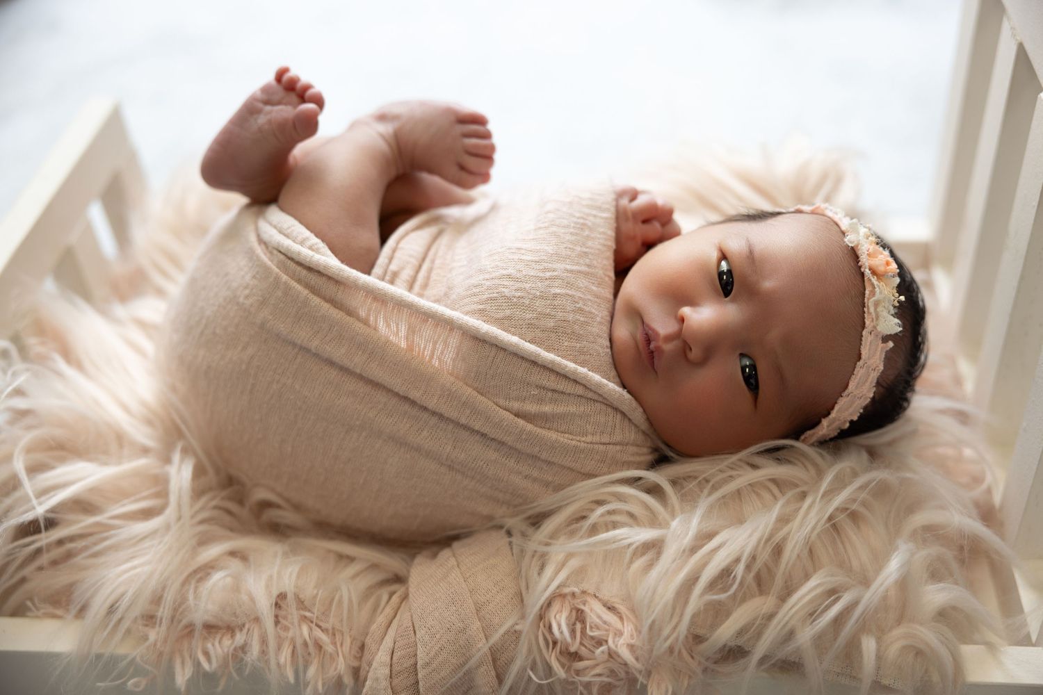 Newborn baby wrapped in beige knit swaddle with floral headband lying on fluffy fur in a white wooden crib.