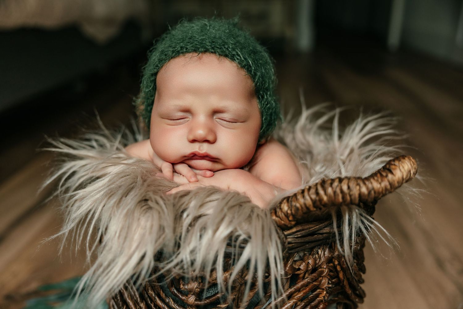 Newborn baby sleeping peacefully in a wicker basket with fur lining, wearing a green knit bonnet hat.