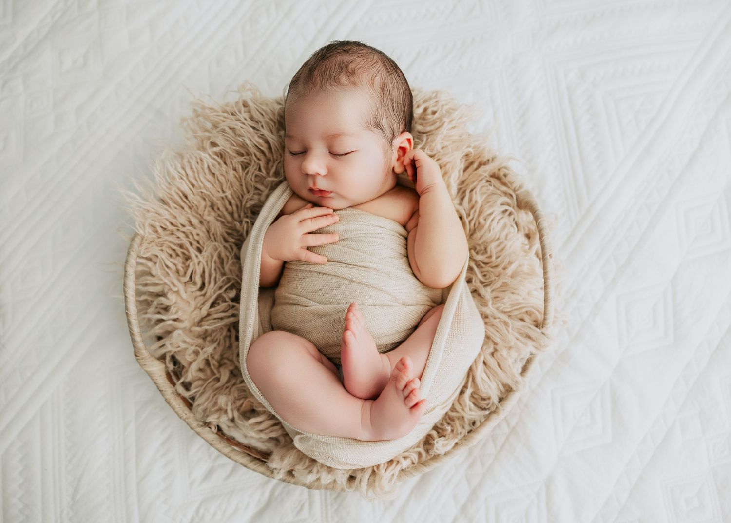 Sleeping newborn baby wrapped in beige swaddle nestled in a round fluffy basket on white background.