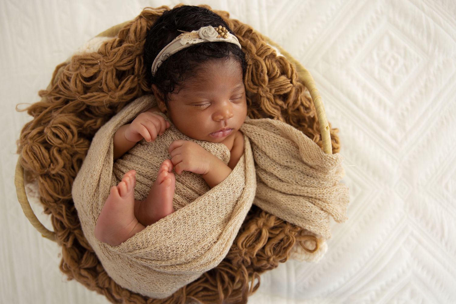 Sleeping newborn baby swaddled in beige wrap with floral headband, nestled in a cozy brown yarn bowl.