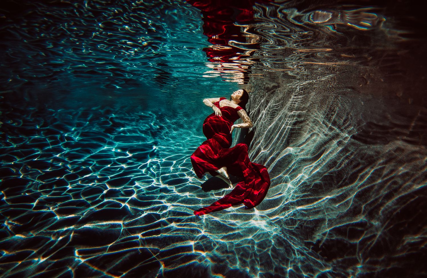 Artistic underwater photography showing a figure in red dress flowing gracefully through dark blue illuminated water.