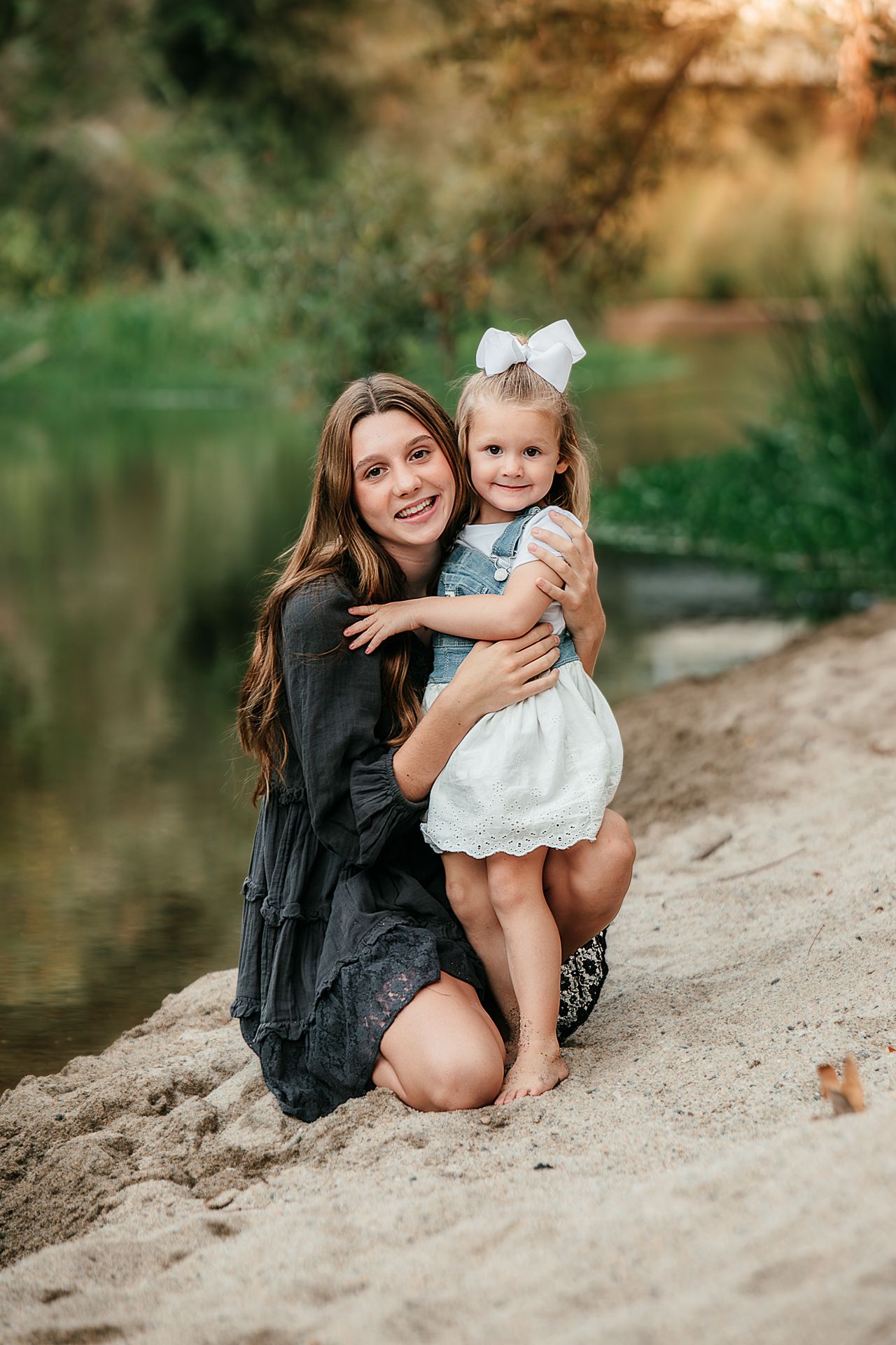 Smiling mother kneeling on sandy riverbank hugging young daughter in white dress and bow in Fallbrook, CA