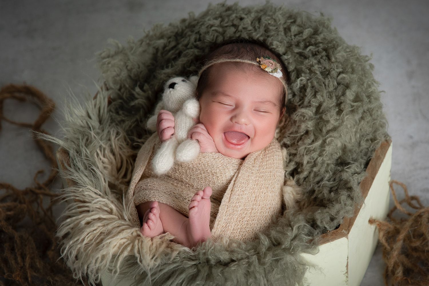 Smiling newborn baby girl in knit wrap holding teddy bear in green fur-lined box.