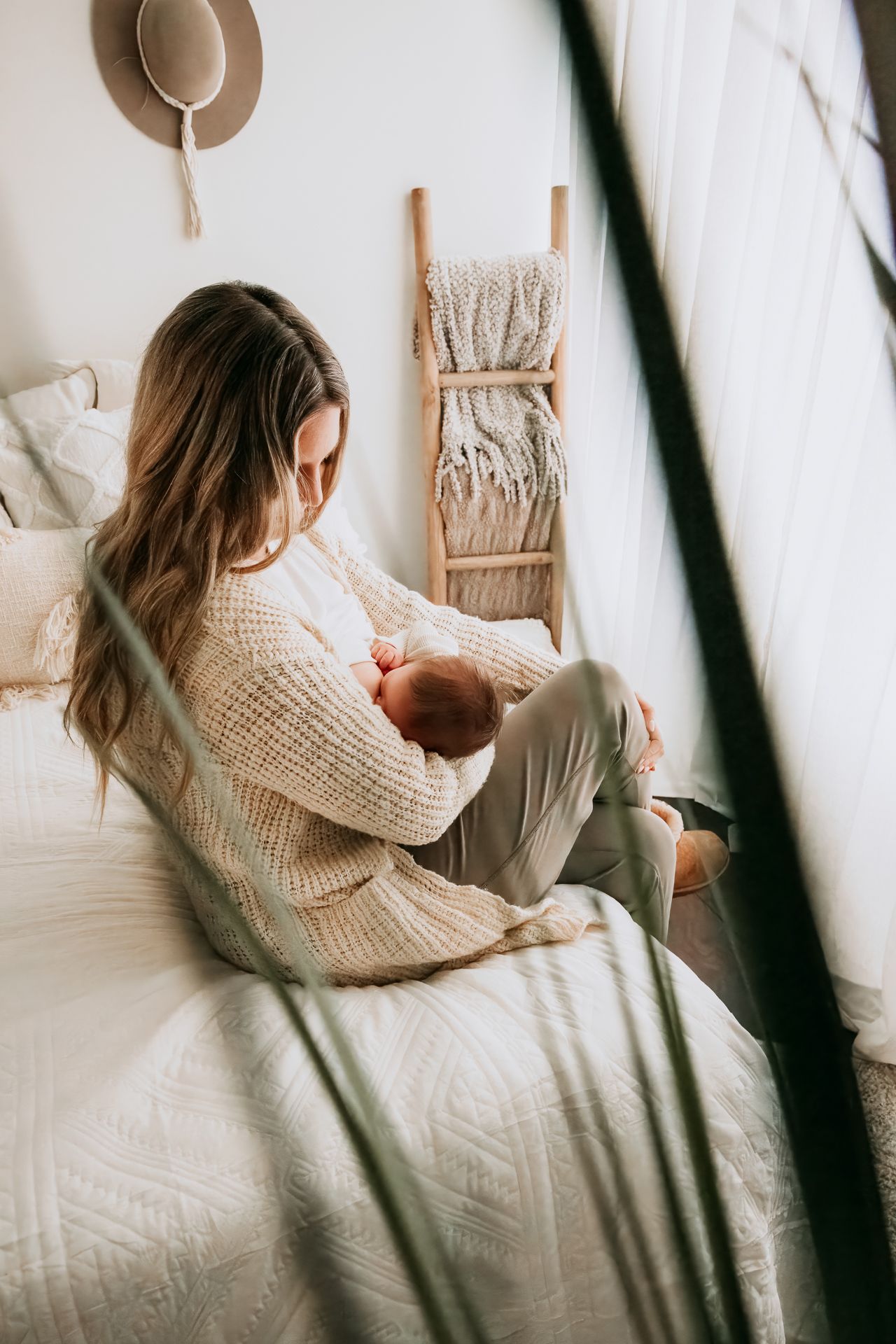 A woman in a cozy knit sweater sits on a bed cradling her newborn baby in a boho-styled bedroom.