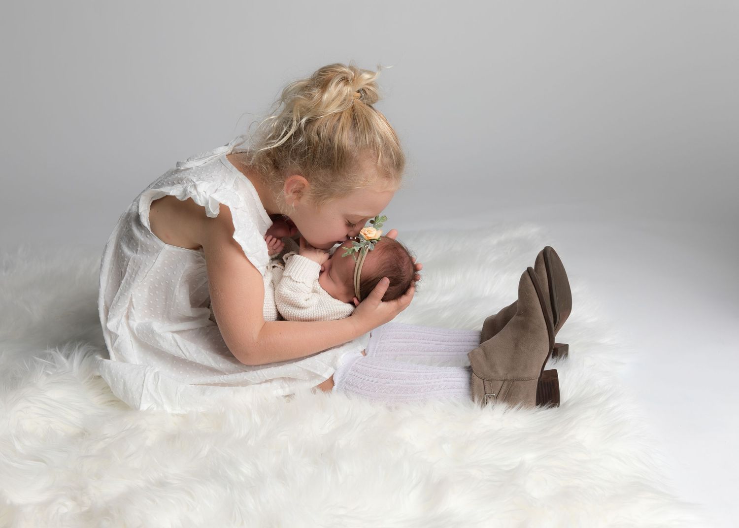 Young girl in white dress kissing newborn baby sibling on fluffy white rug in professional studio photo.
