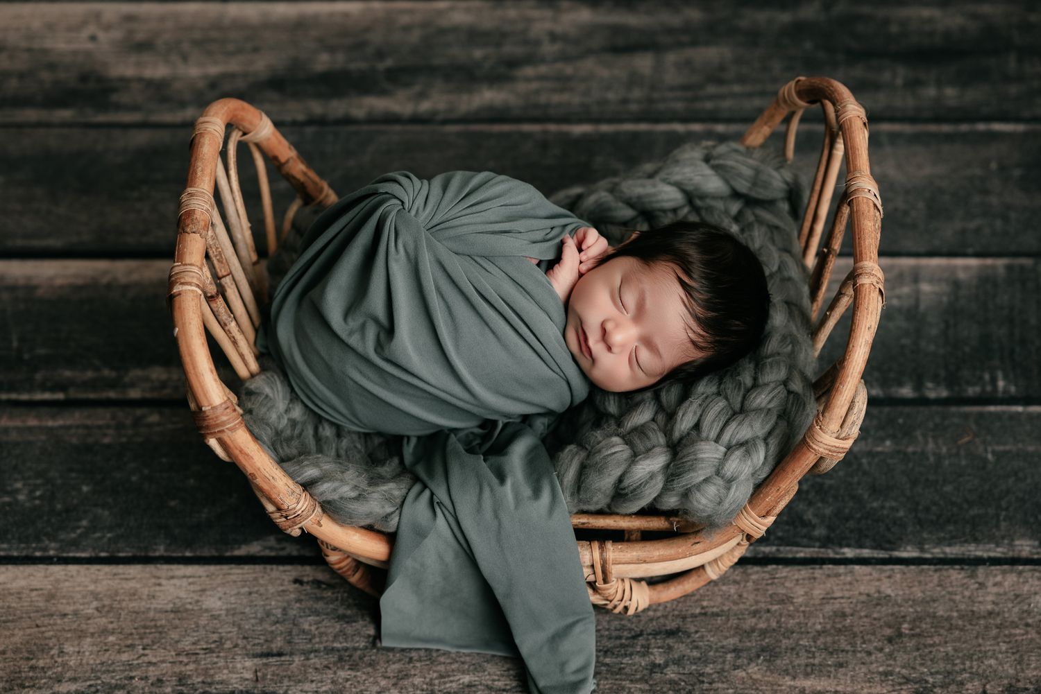Newborn baby swaddled in sage green wrap sleeping peacefully in a rustic wicker basket with chunky knit blanket.