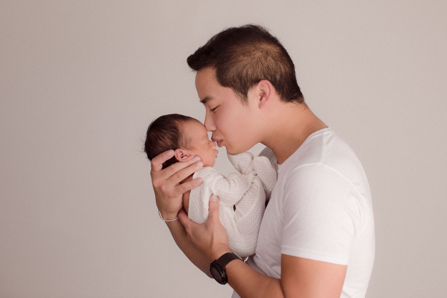 A father tenderly kisses his newborn baby while cradling the infant in both hands against a neutral background.