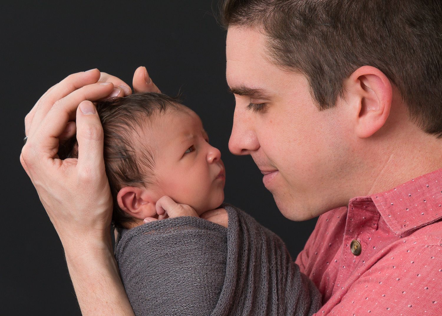 A father tenderly holds his newborn baby wrapped in gray, gazing lovingly at the infant against a dark background.