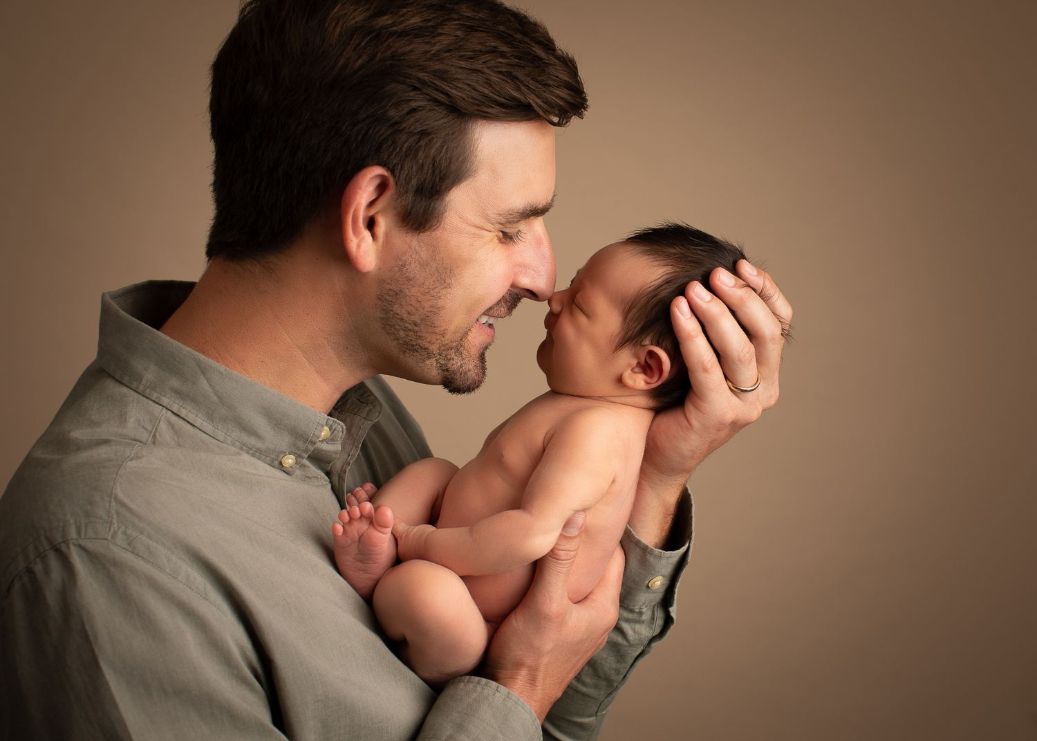 Father tenderly holding newborn baby close, sharing an intimate nose-to-nose moment against a warm beige background.