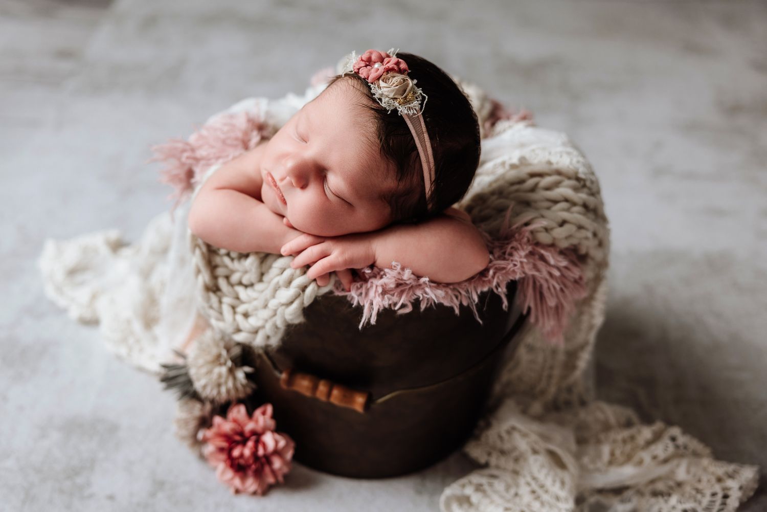 Sleeping newborn baby girl in a wooden bucket with pink floral headband and soft knit wrap.