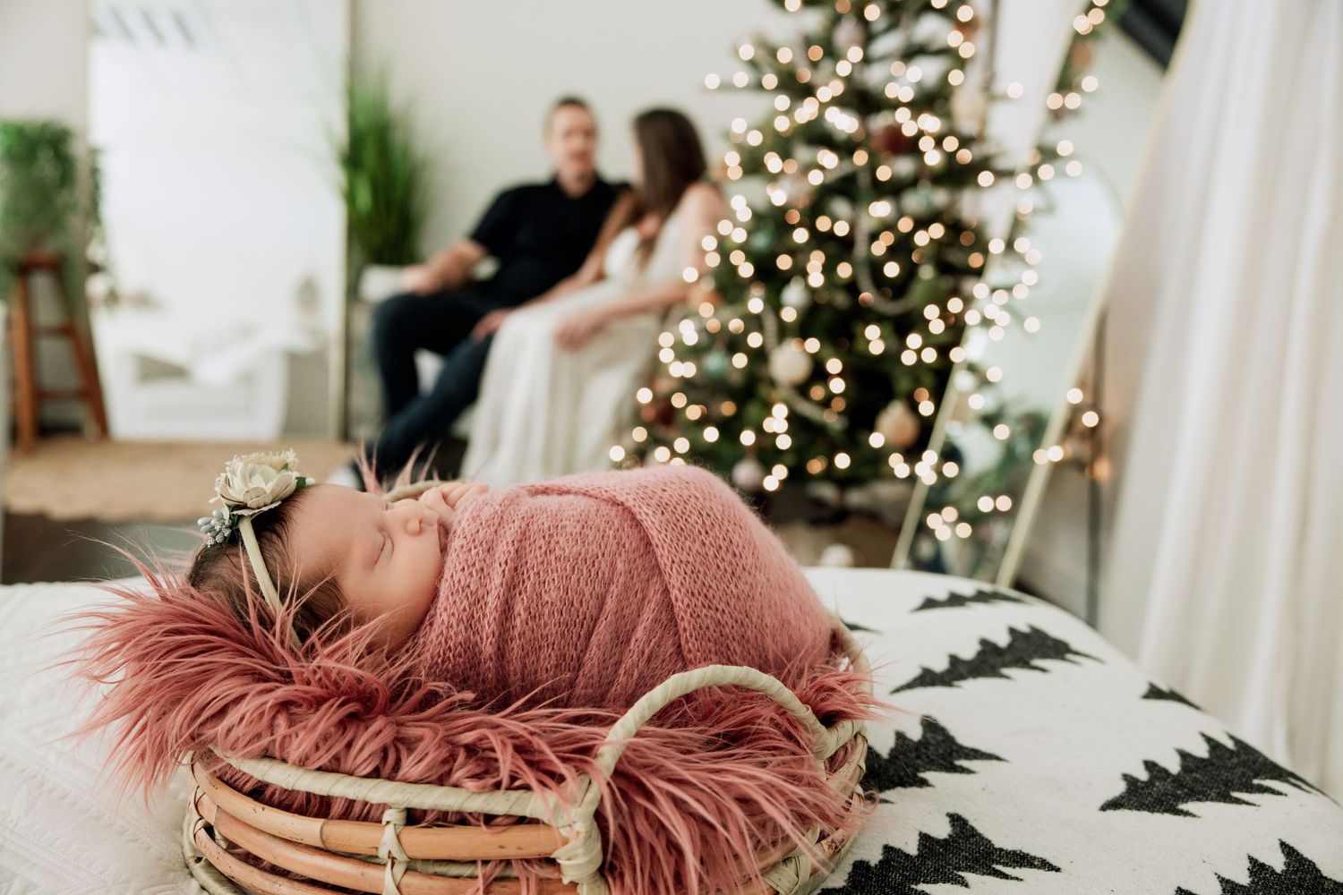 Newborn baby wrapped in pink in a basket with fur, parents and Christmas tree softly blurred in background.