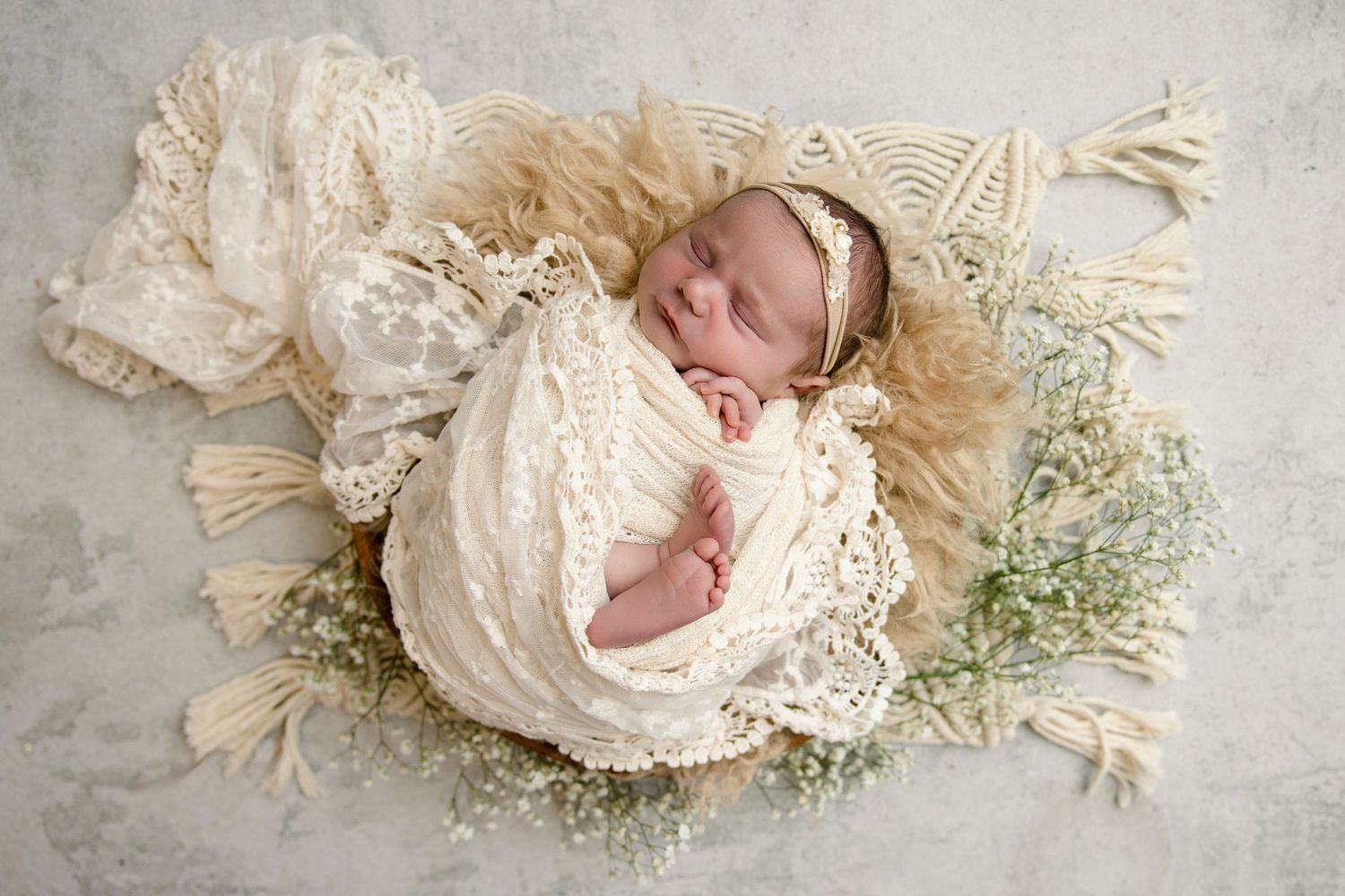 Peaceful newborn wrapped in ivory lace swaddle with headband, nestled in basket with baby's breath on cream macrame blanket.