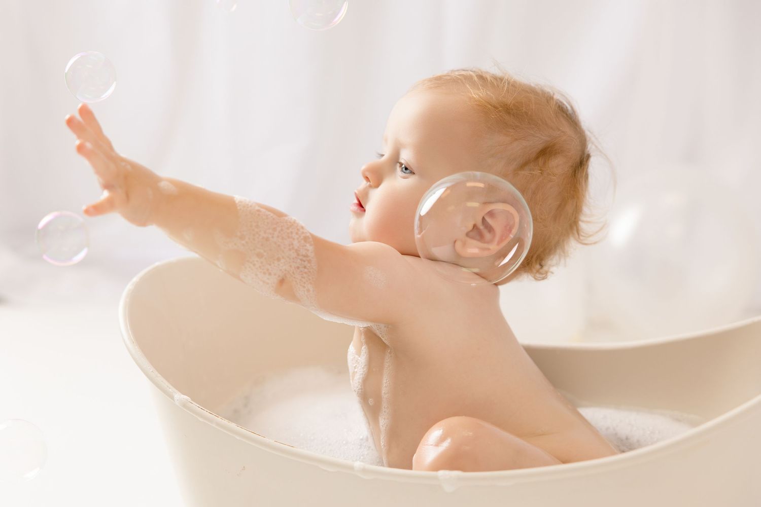 Cute baby sitting in a white bathtub with bubbles, reaching out to touch soap bubbles during bath time.