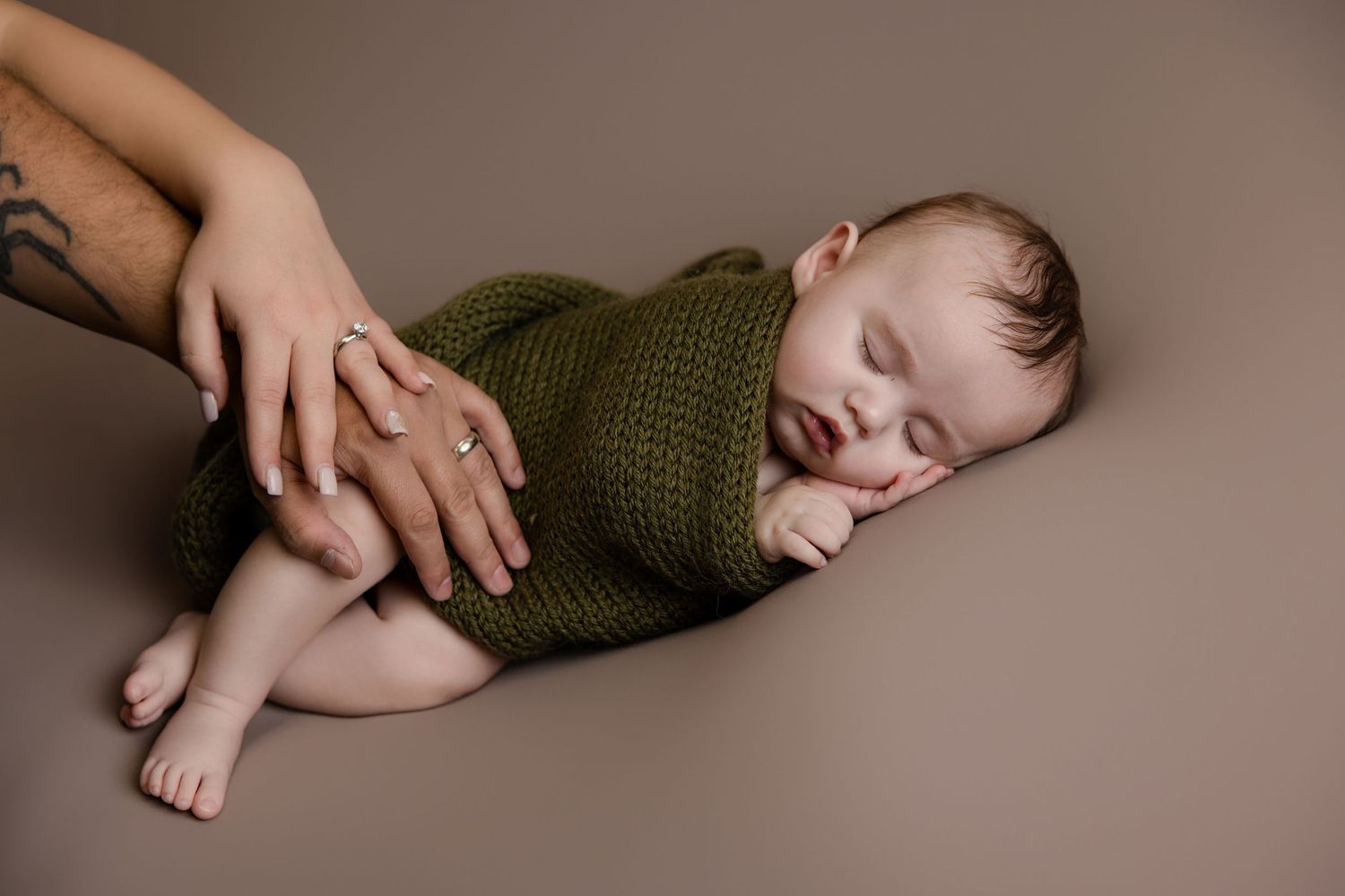 Sleeping newborn baby wrapped in green knit blanket with parent's tattooed hands gently cradling on beige background.