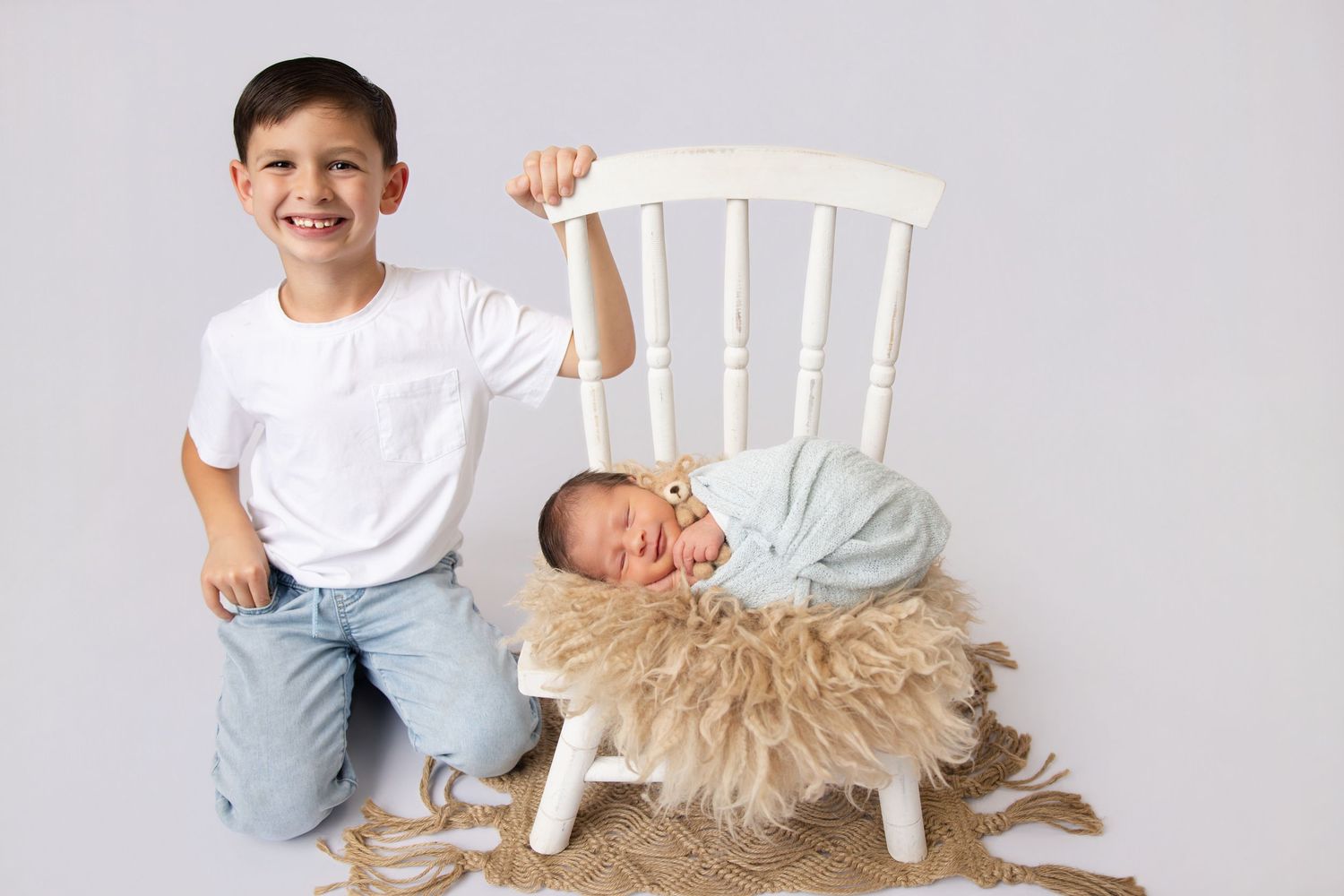 Smiling boy kneels beside a white chair holding a sleeping newborn in a cozy fur-lined basket.