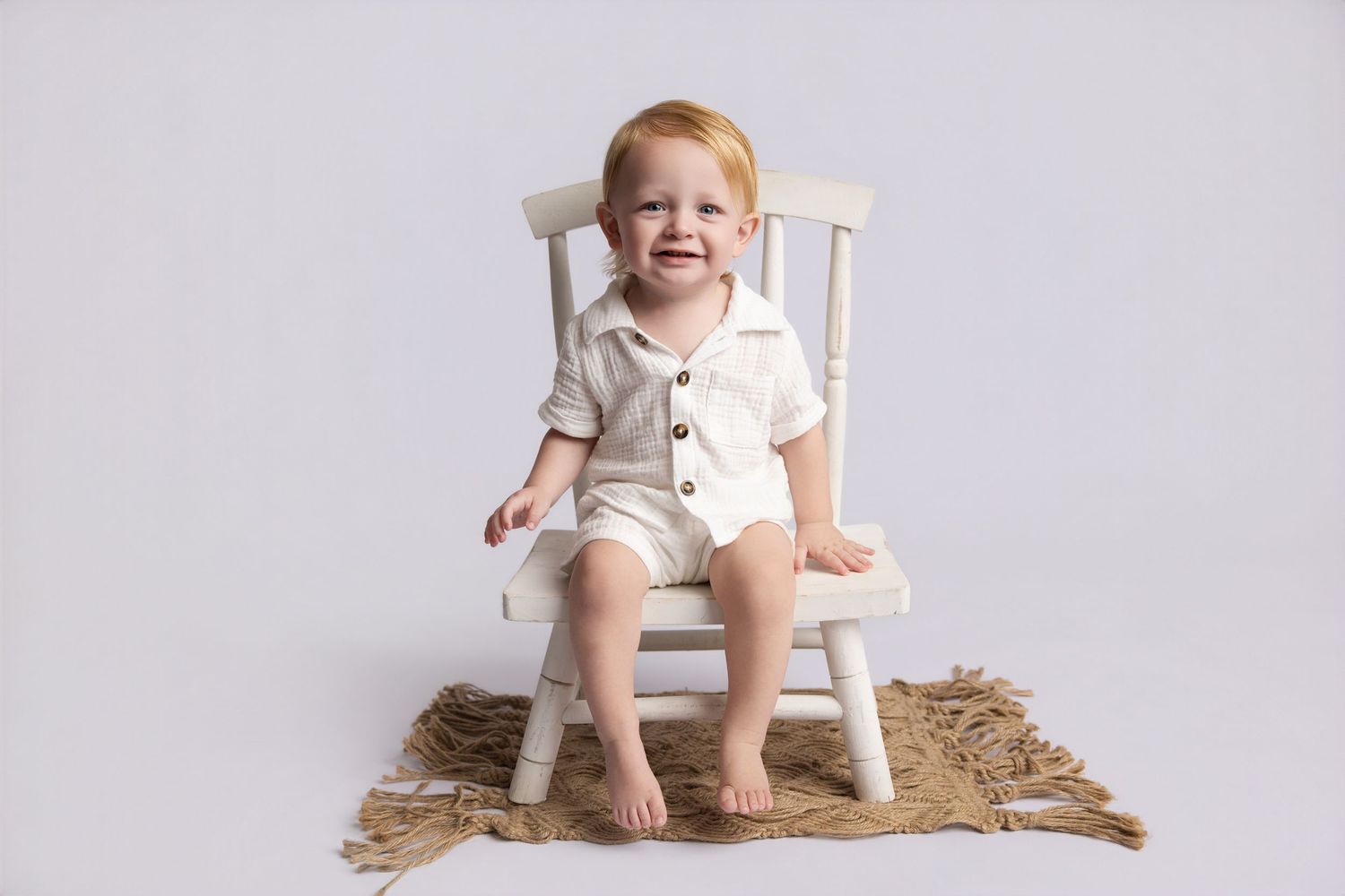 Smiling toddler boy in white romper sitting on small white chair on burlap rug against gray background.