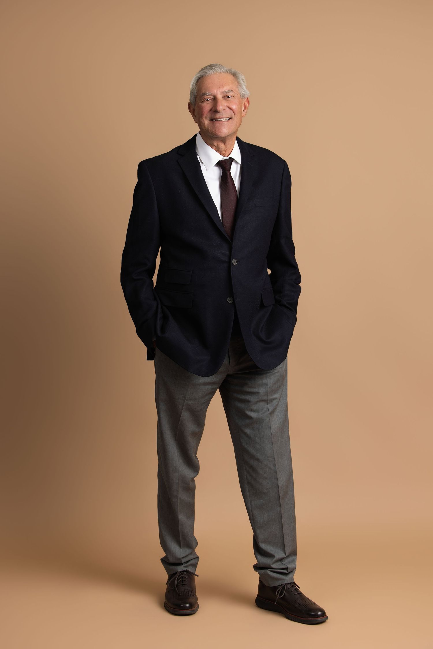 Smiling senior businessman in navy blazer, white shirt, and tie with hands in pockets on beige background.