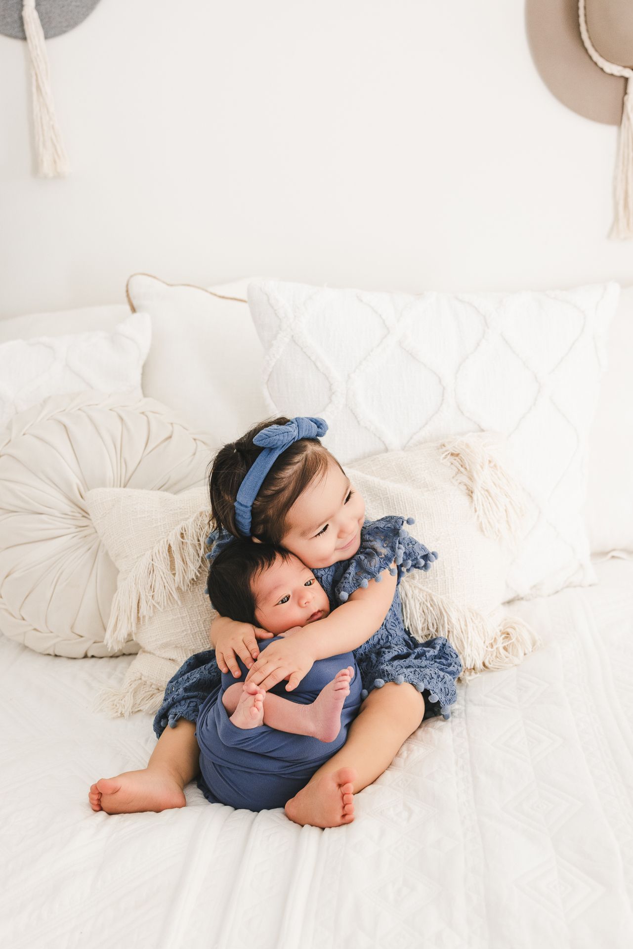 Young girl in blue dress hugging newborn sibling on white bed with decorative pillows in bright nursery setting.