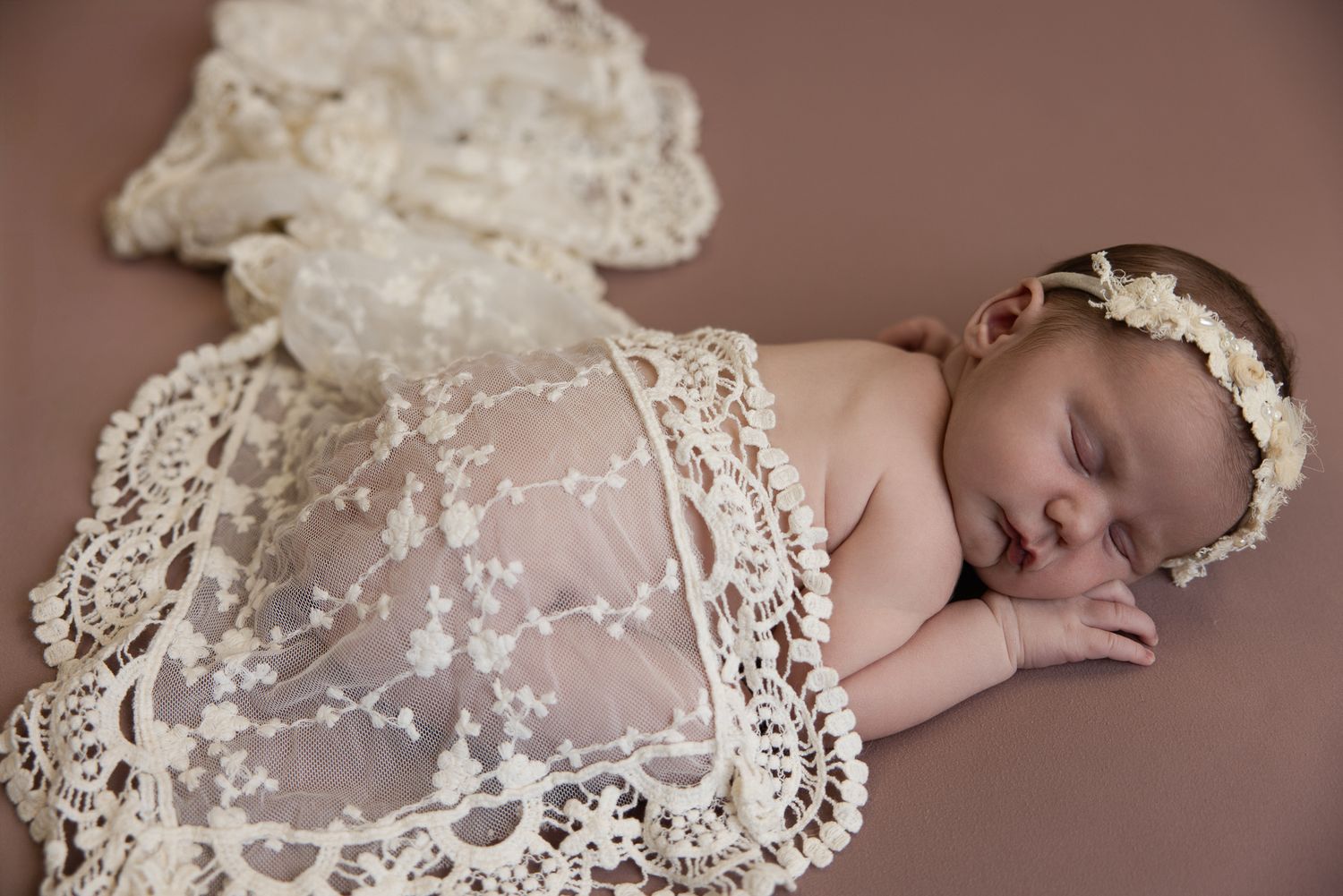 Sleeping newborn baby wrapped in delicate ivory lace fabric wearing a floral headband on a brown backdrop.
