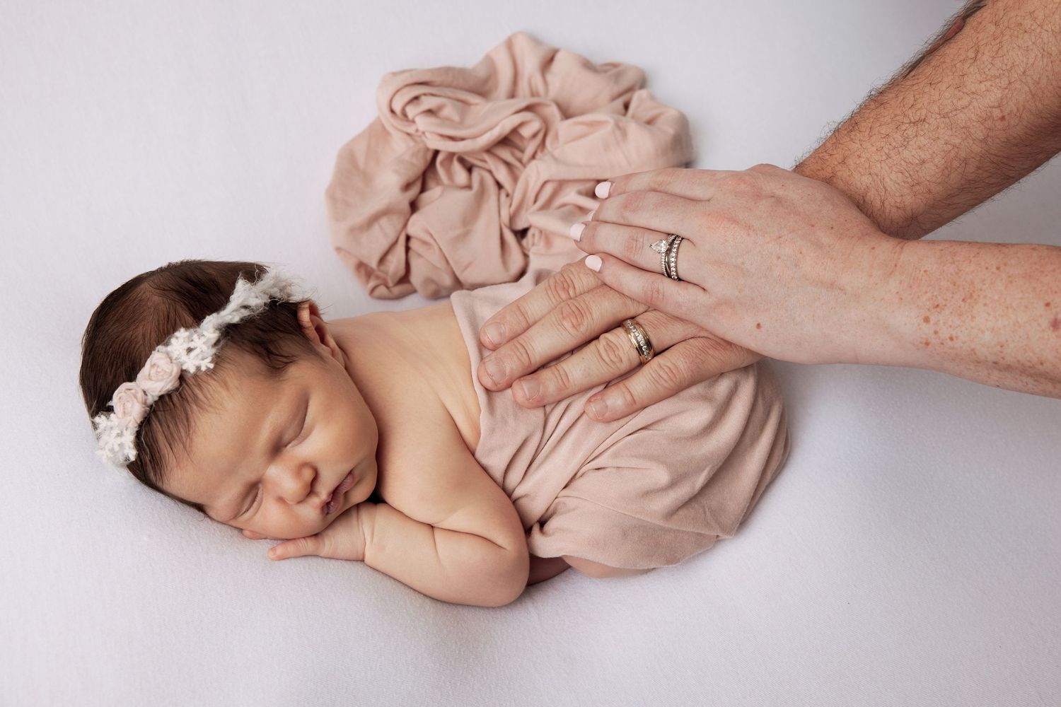 Sleeping newborn baby girl in pink wrap with floral headband with parent hands gently resting on her back.