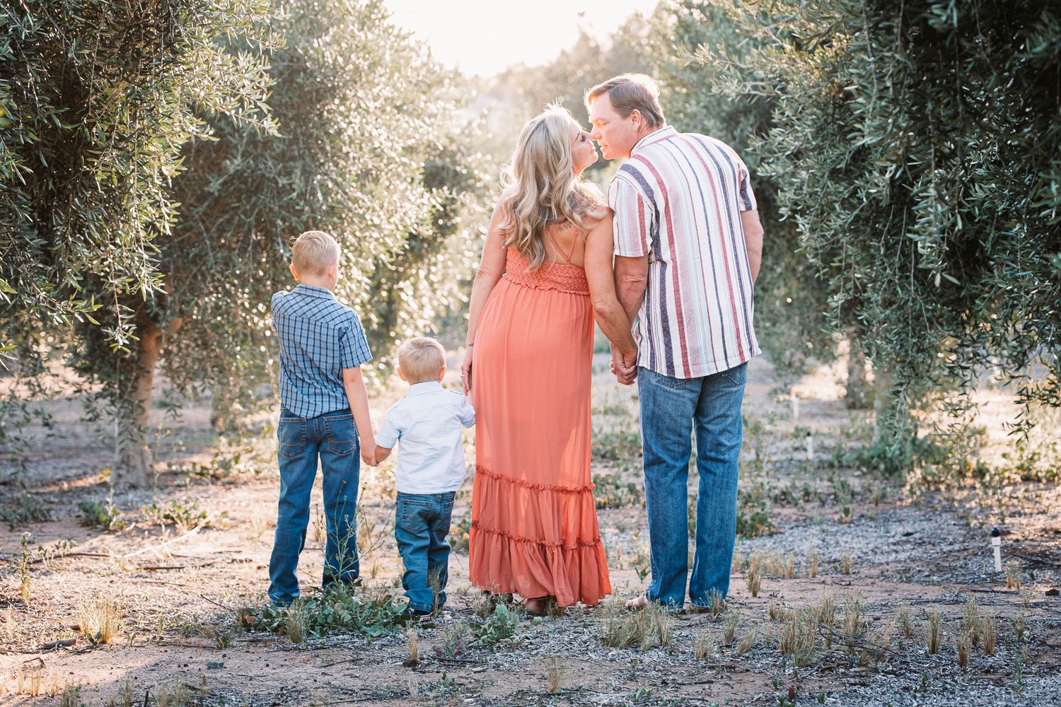 Family of four holding hands from behind in an olive grove during golden hour in Bonsall, near Fallbrook, & Temecula