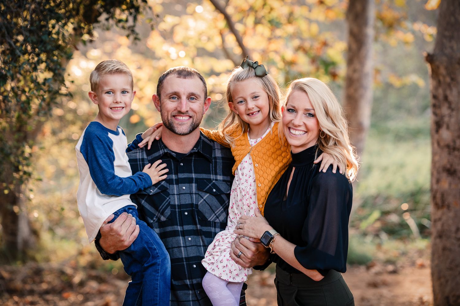Smiling family of four poses together among golden fall trees during outdoor portrait session at a Preserve in Fallbrook