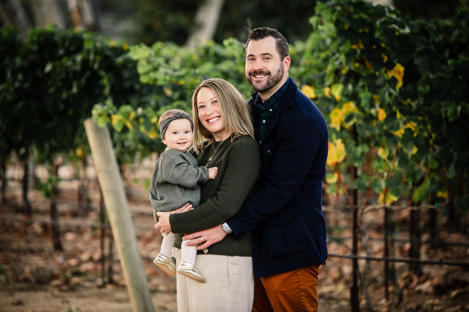 Smiling couple holding baby in a lush vineyard setting.
