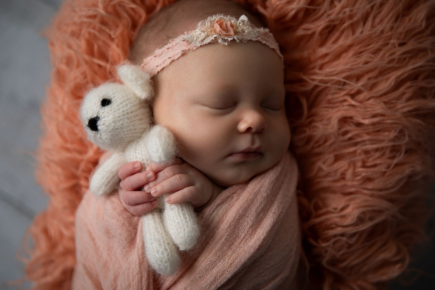 Peaceful newborn baby girl in pink wrap holding a knit teddy bear on a soft peach blanket.