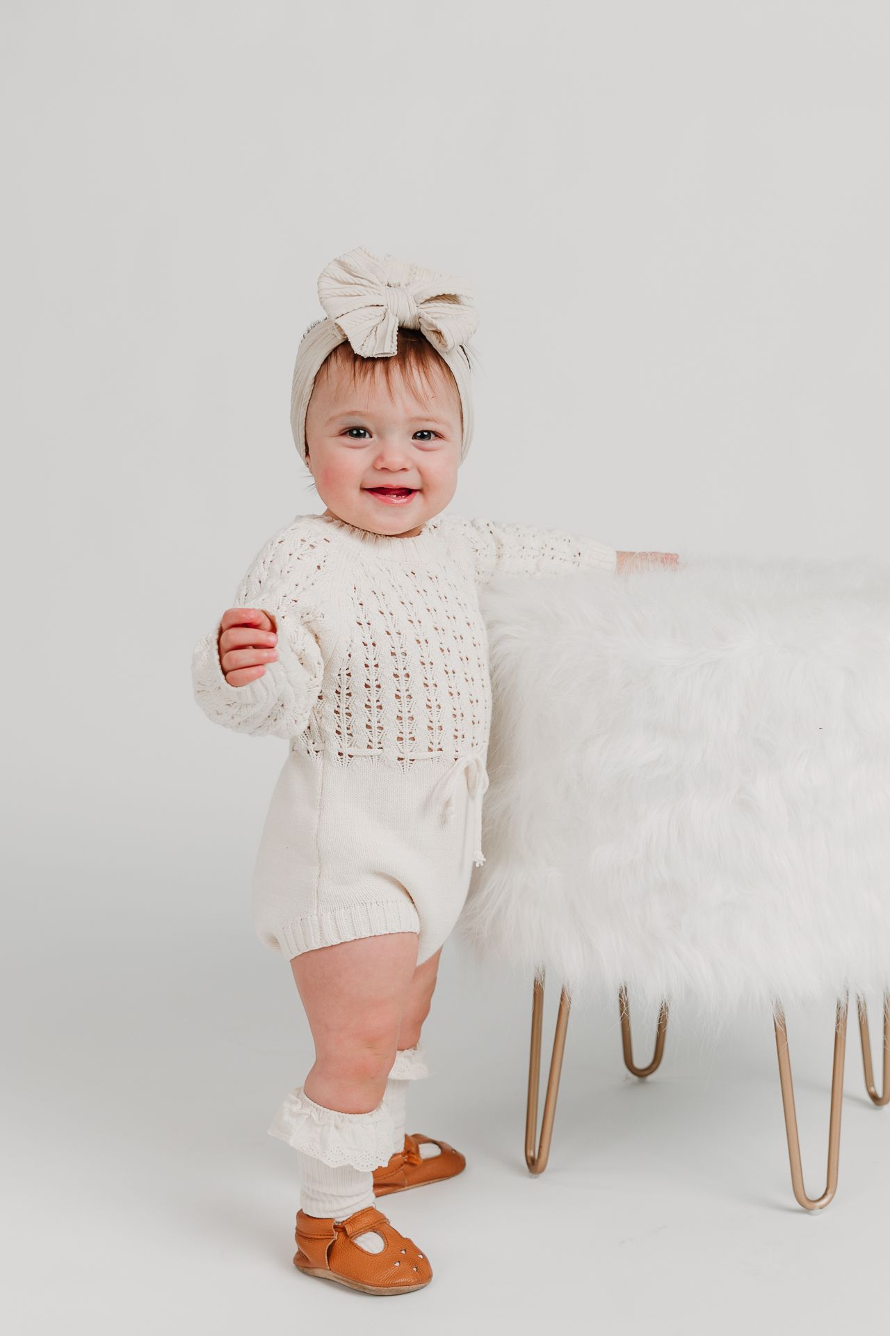 Happy baby girl in white knit outfit and bow headband standing beside a fluffy white ottoman.