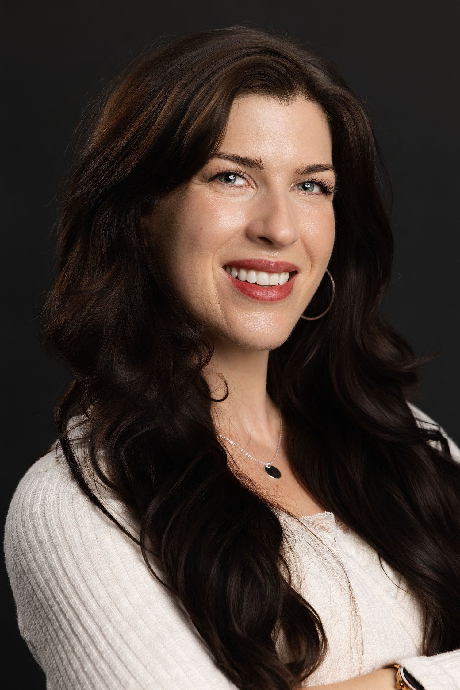 Smiling woman with long dark wavy hair wearing a white sweater and hoop earrings against a dark background.
