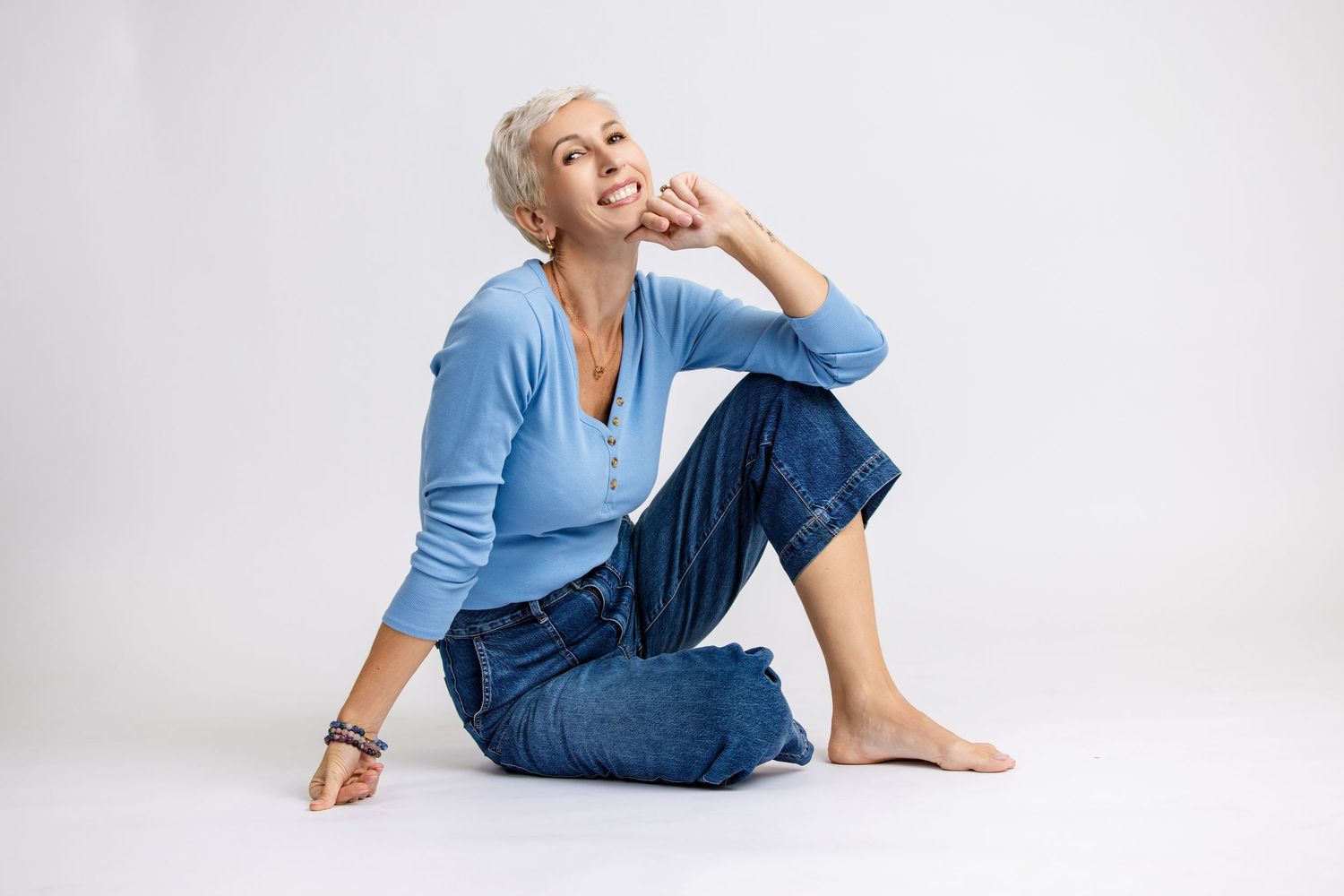 Mature woman with short gray hair sitting barefoot on floor in blue top and jeans, smiling thoughtfully.