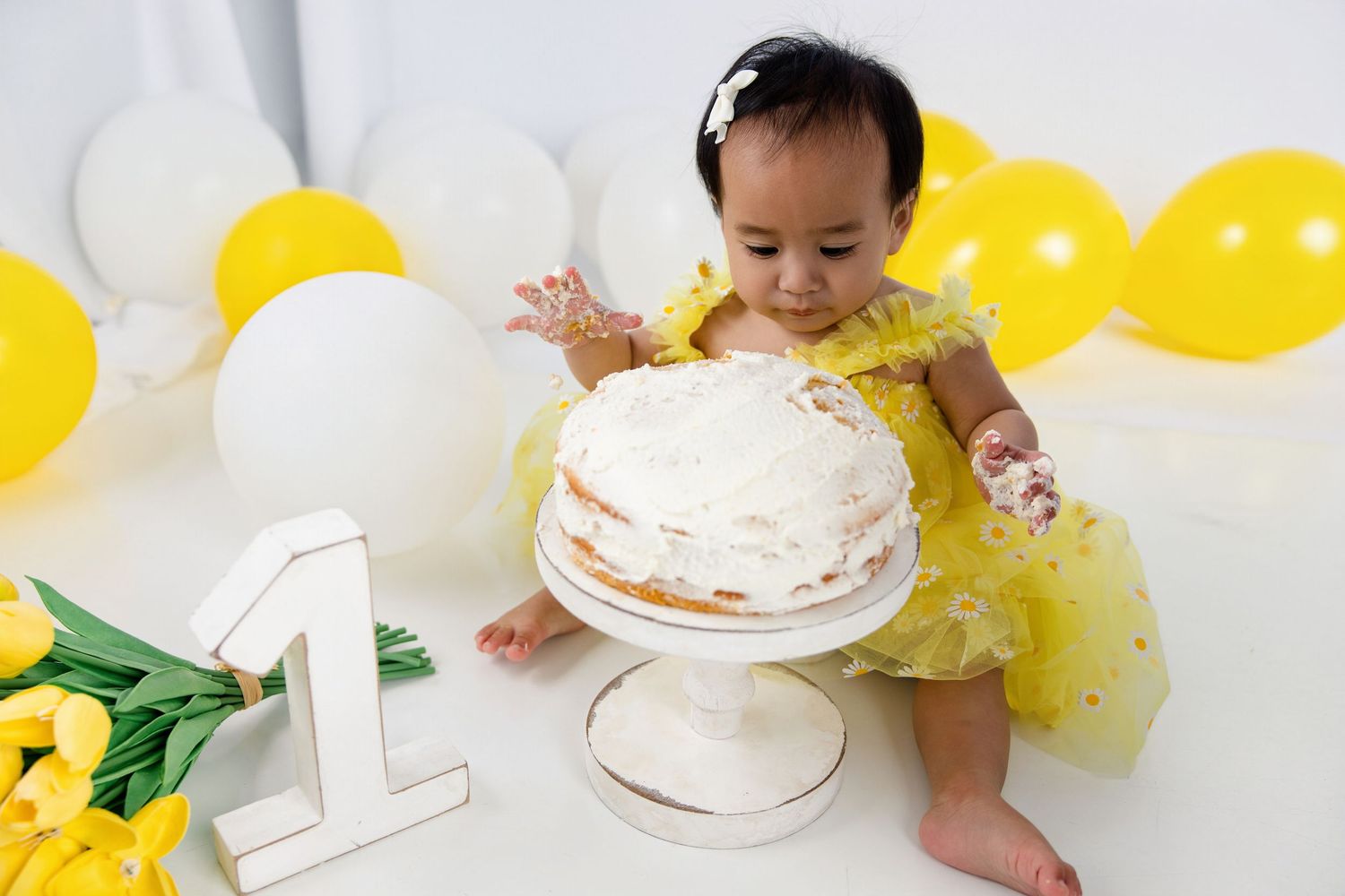 Baby girl in yellow dress smashing her first birthday cake surrounded by yellow balloons and a number one decoration.