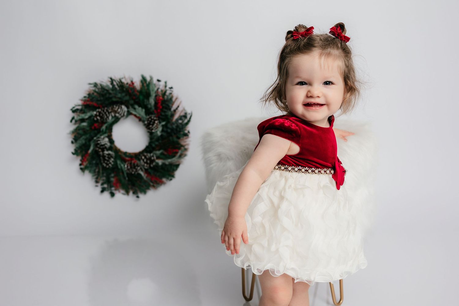 Toddler girl in red and white Christmas dress with bows smiling near holiday wreath on white background.