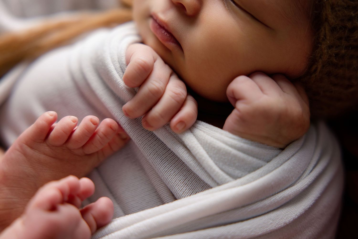 Close-up of a sleeping newborn baby swaddled in a white wrap, with tiny hands tucked under chin.