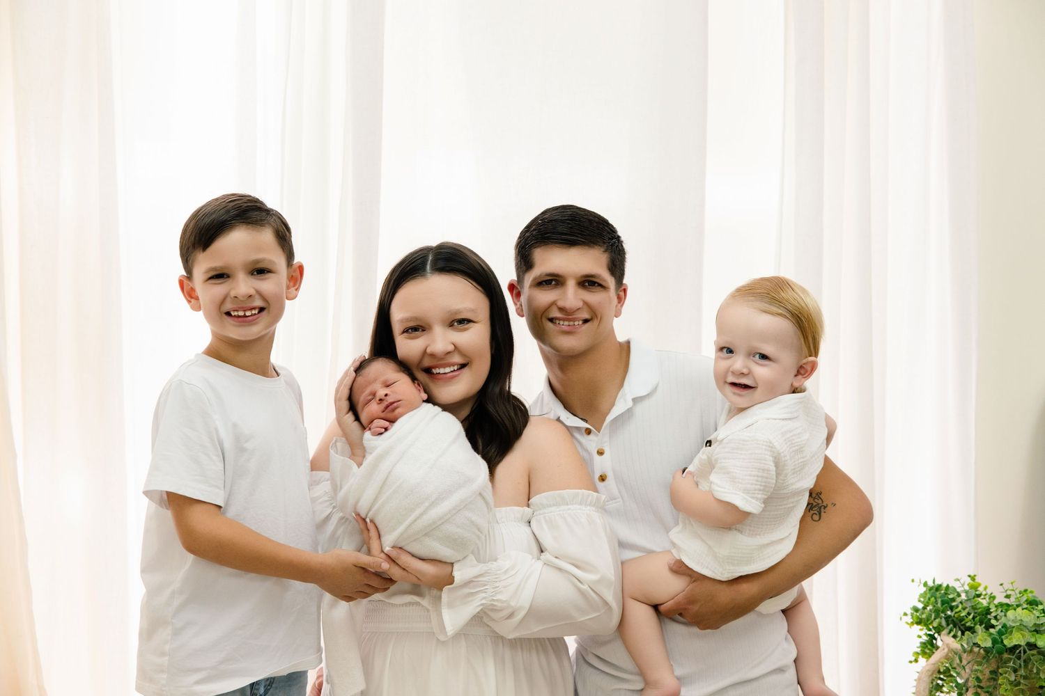 Happy family of five posing together, parents holding three young children including a newborn baby, in a bright white room.