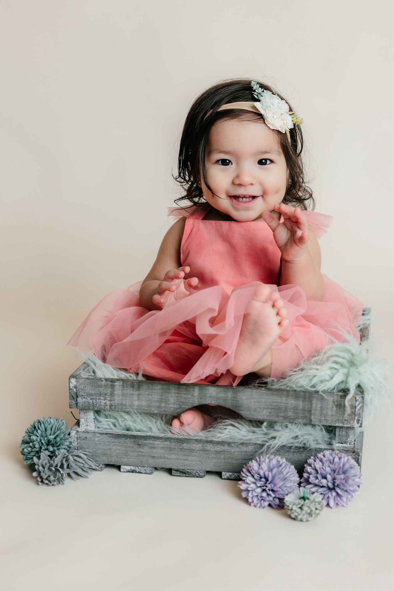 Smiling baby girl in pink tutu dress sitting in rustic wooden crate with purple flowers and floral headband.