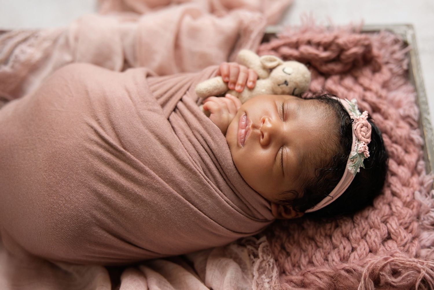 Newborn baby girl swaddled in pink wrap, wearing floral headband, sleeping peacefully on a soft pink blanket.