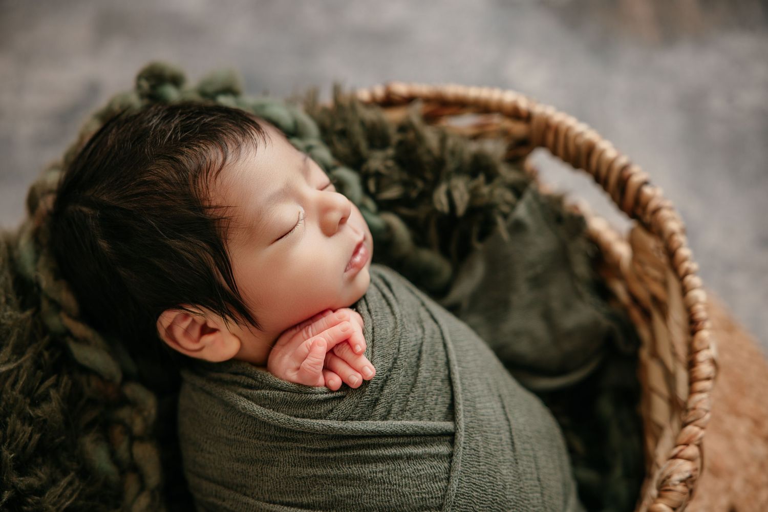 Peaceful newborn baby swaddled in green wrap sleeping in a wicker basket with soft green blanket.