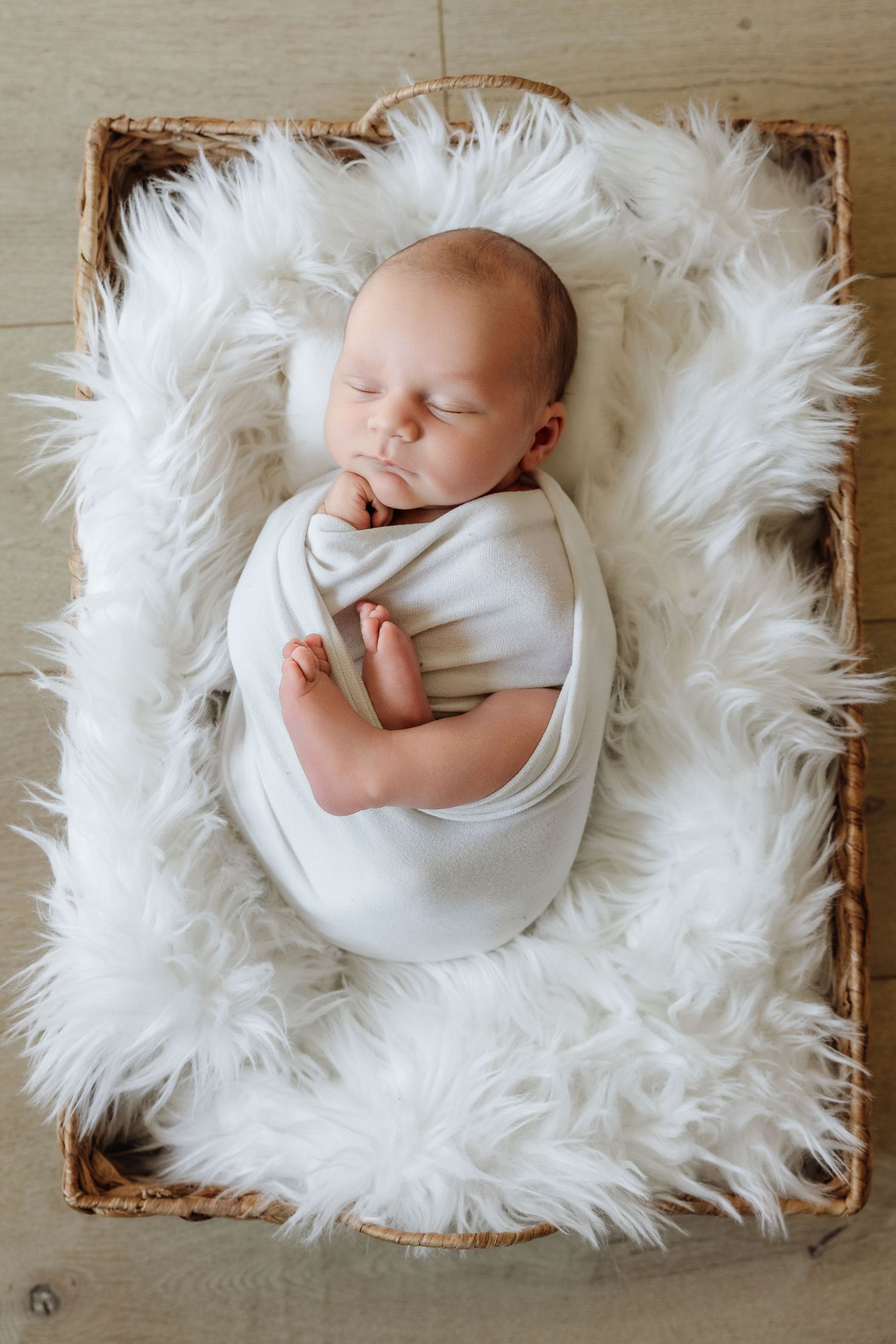 Peaceful newborn baby swaddled in white, sleeping in a wicker basket lined with fluffy white faux fur.