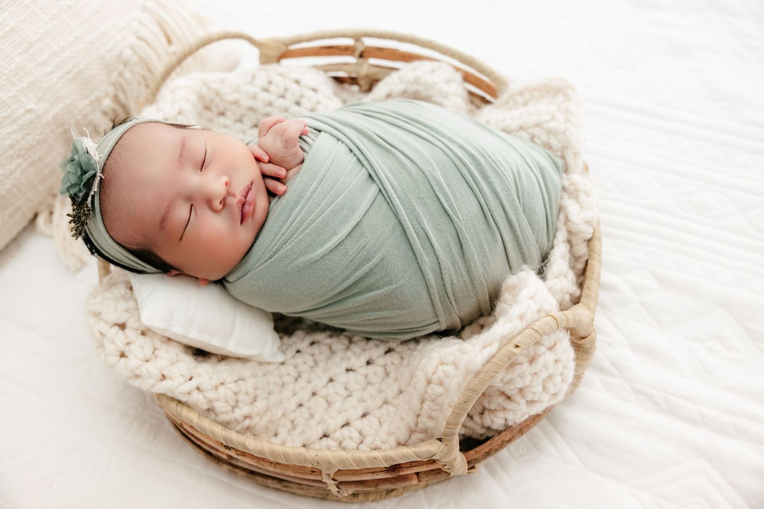 Sleeping newborn baby swaddled in sage green wrap with floral headband, resting in a wicker basket.