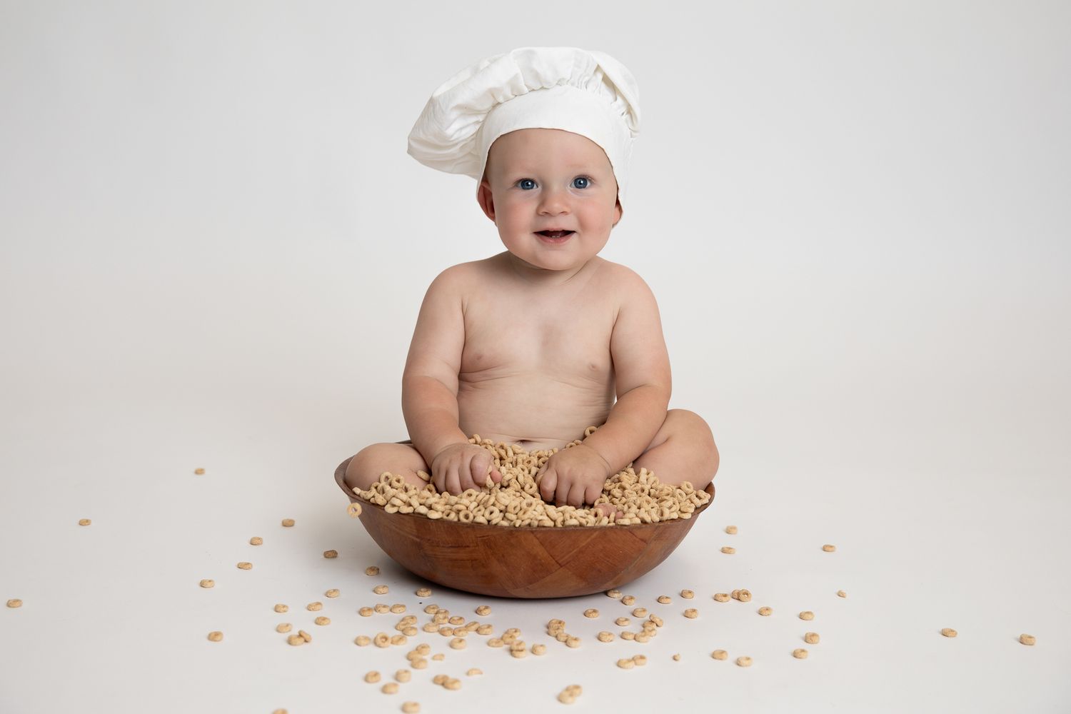 Smiling baby in white chef hat sitting inside a large wooden bowl filled with puffed cereals.