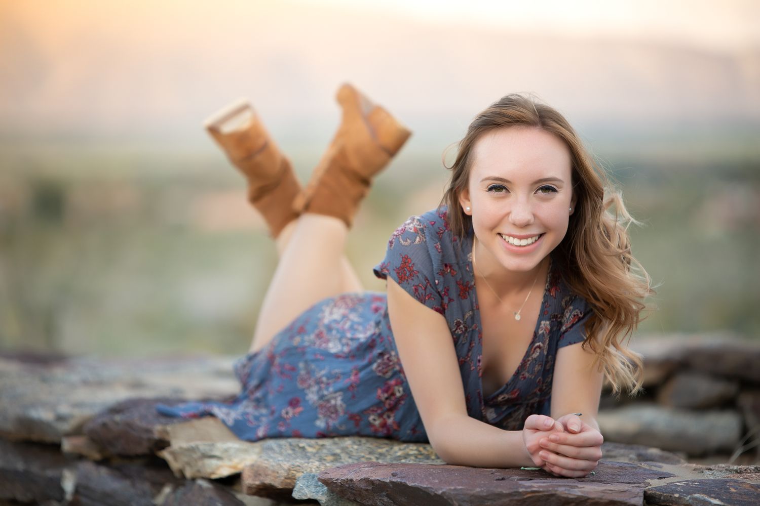 Smiling young woman in floral dress lies on stone wall with boots up at sunset.