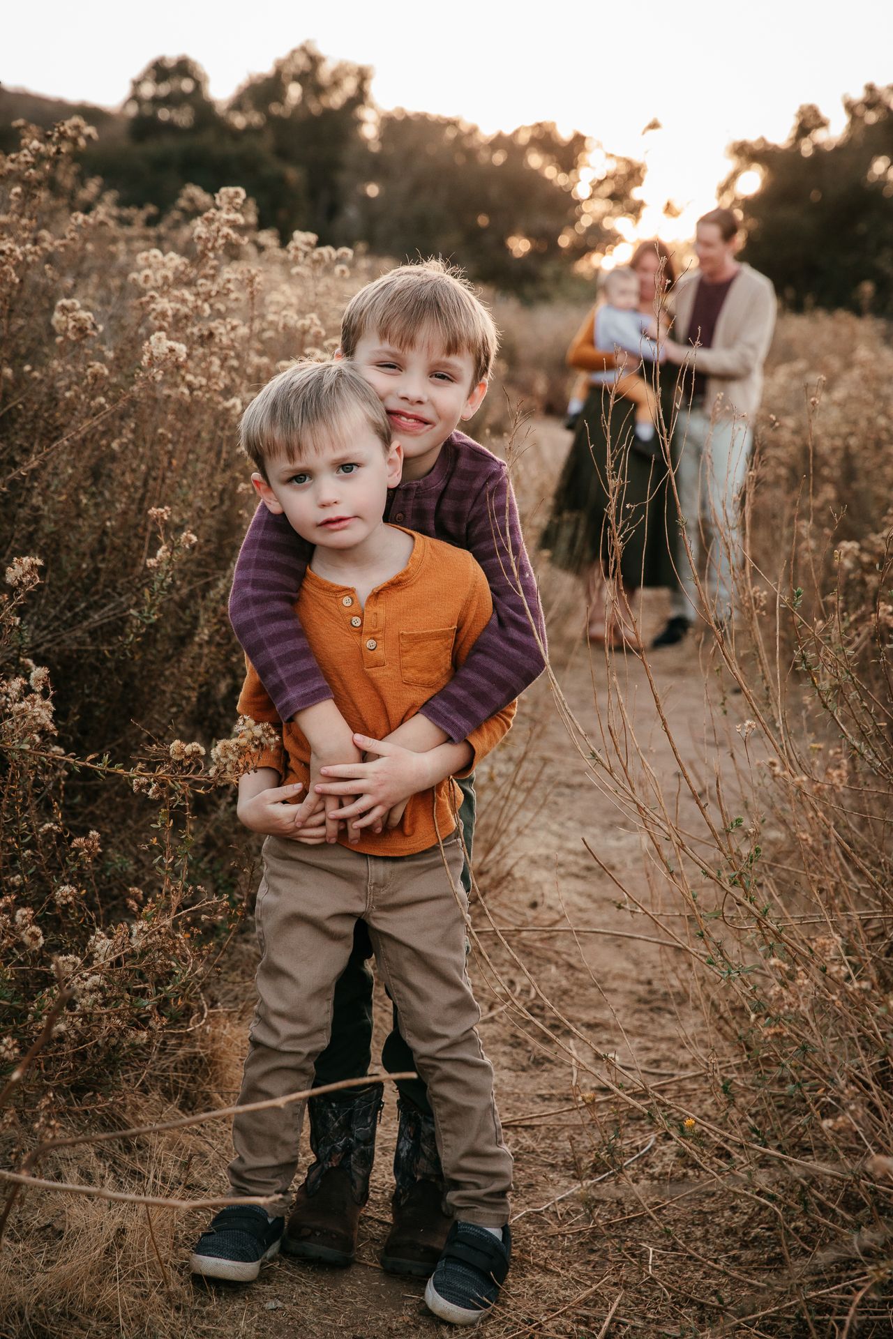 Two young brothers hugging in wildflower field with parents embracing softly in background.