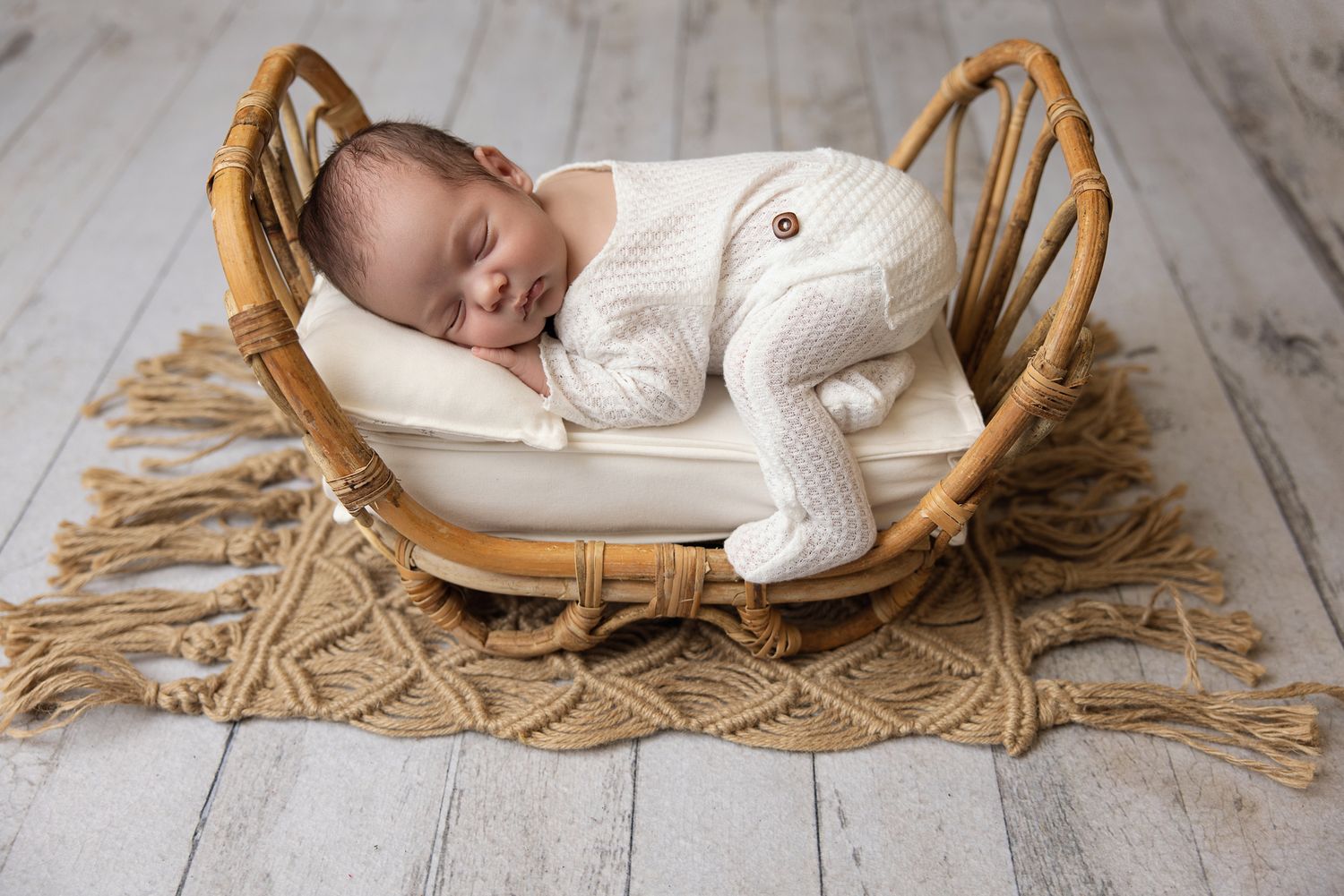 Newborn baby sleeping peacefully in a wicker bassinet wearing a white knit outfit on a rustic wood floor.