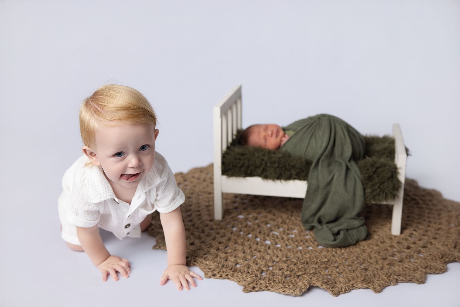 Toddler boy smiling on floor beside newborn baby sleeping in small white bed wrapped in green swaddle.