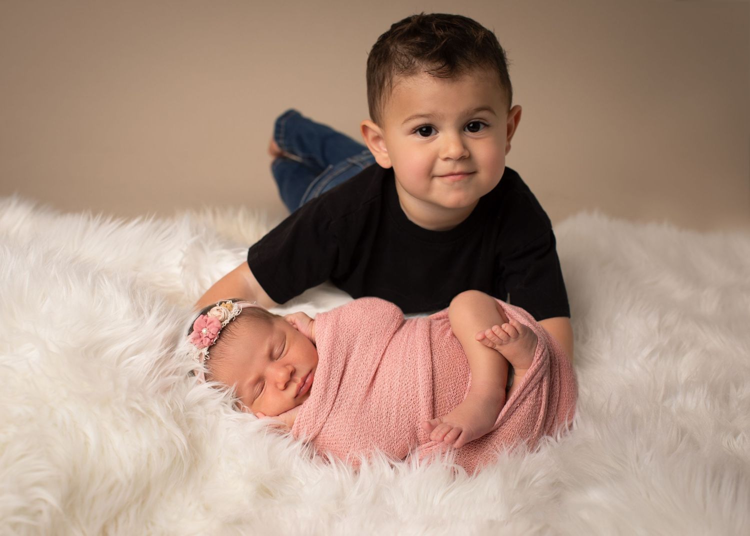 Toddler boy in black shirt lying behind sleeping newborn baby girl wrapped in pink on white fur blanket.