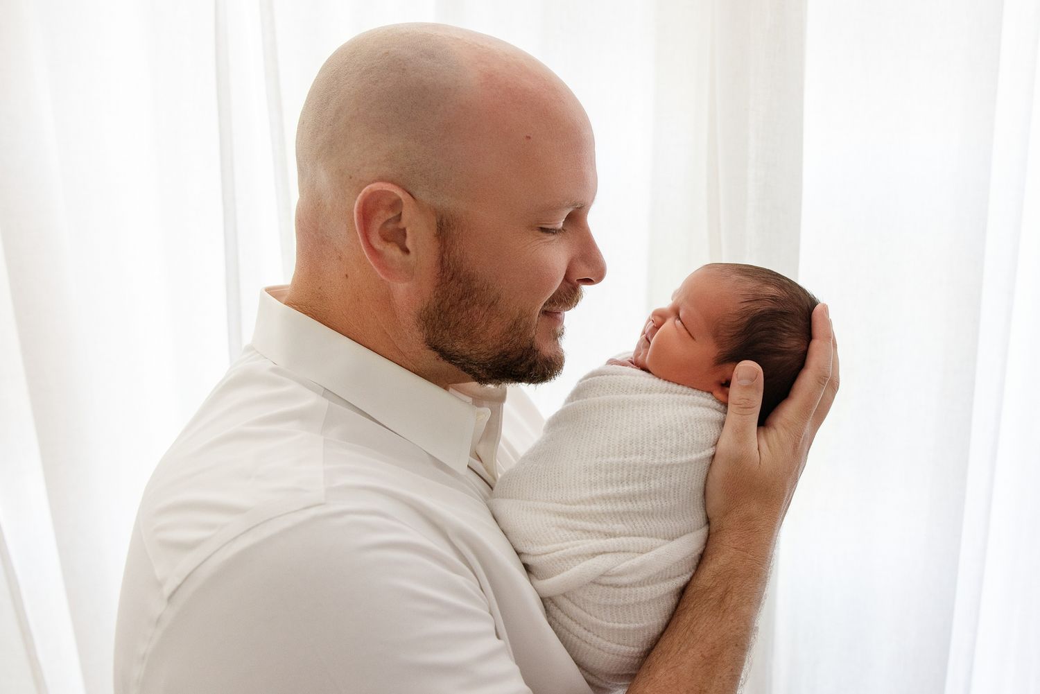 Bald father tenderly holds sleeping newborn baby wrapped in white swaddle near bright window.