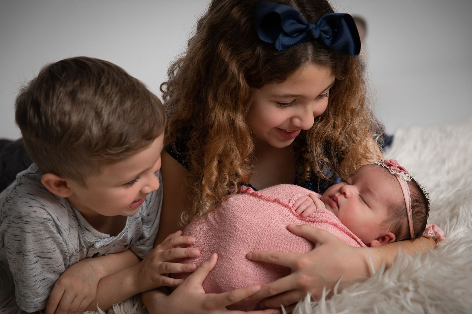 Two older siblings lovingly hold their newborn baby wrapped in pink on a soft white blanket.