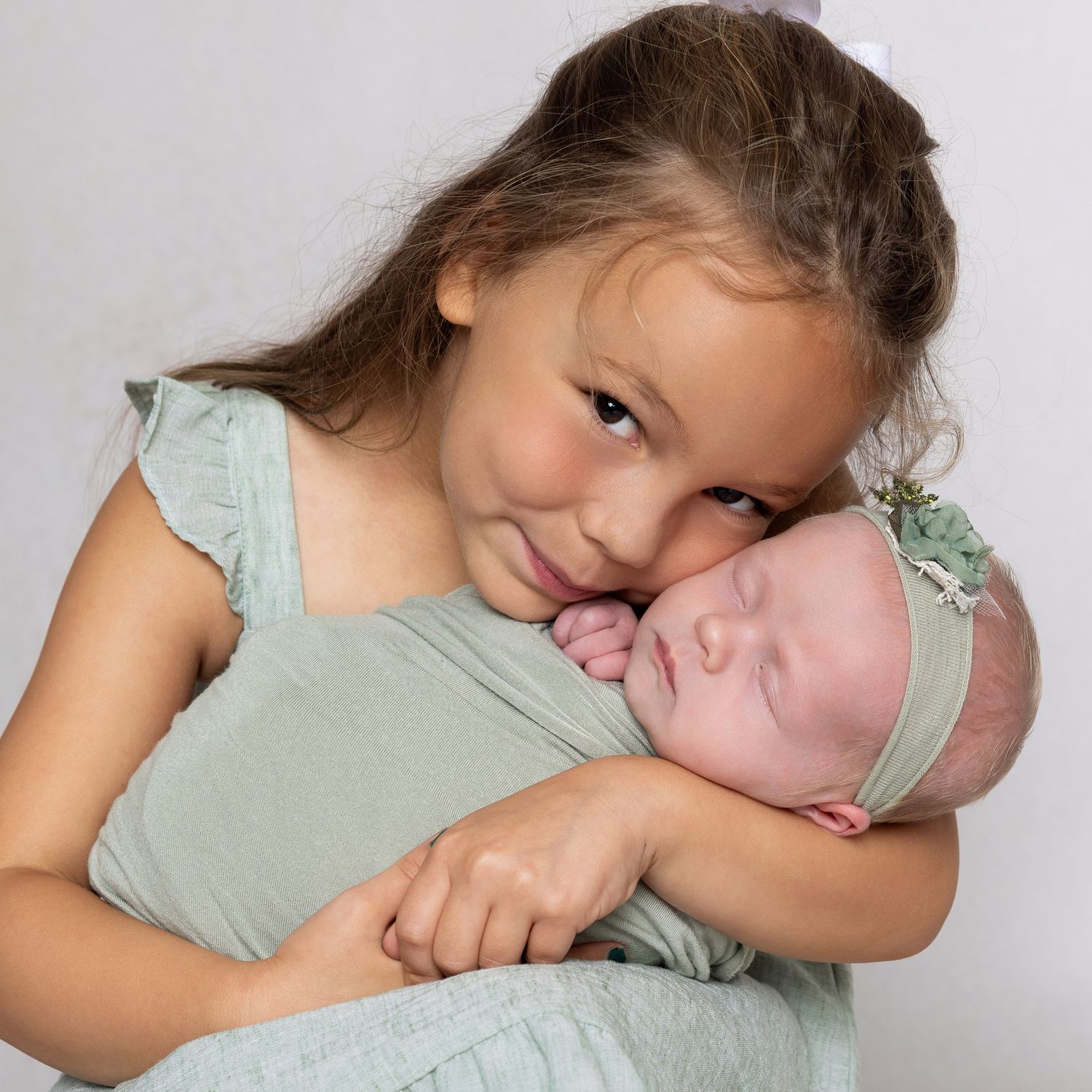 A young girl in a mint dress tenderly holds a sleeping newborn baby wrapped in a matching green swaddle.