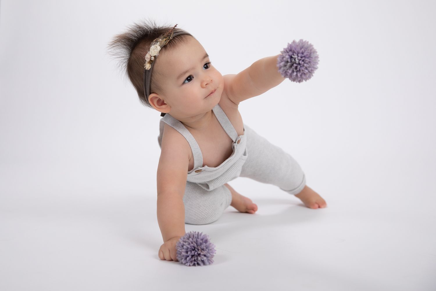 Baby girl in gray romper crawling with purple pom pom toys on white backdrop.